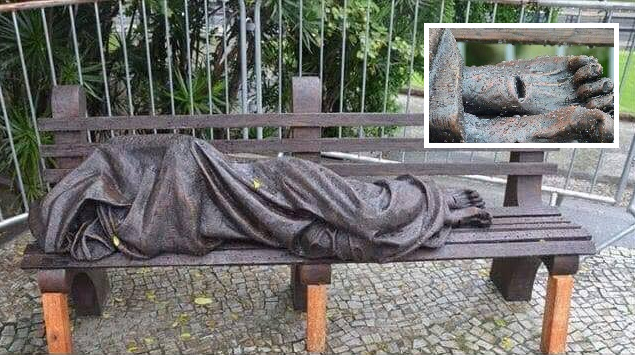 Fotografia da escultura Jesus Sem-Teto, de Timothy P. Schmalz: uma figura coberta sobre um banco, com detalhe dos pés